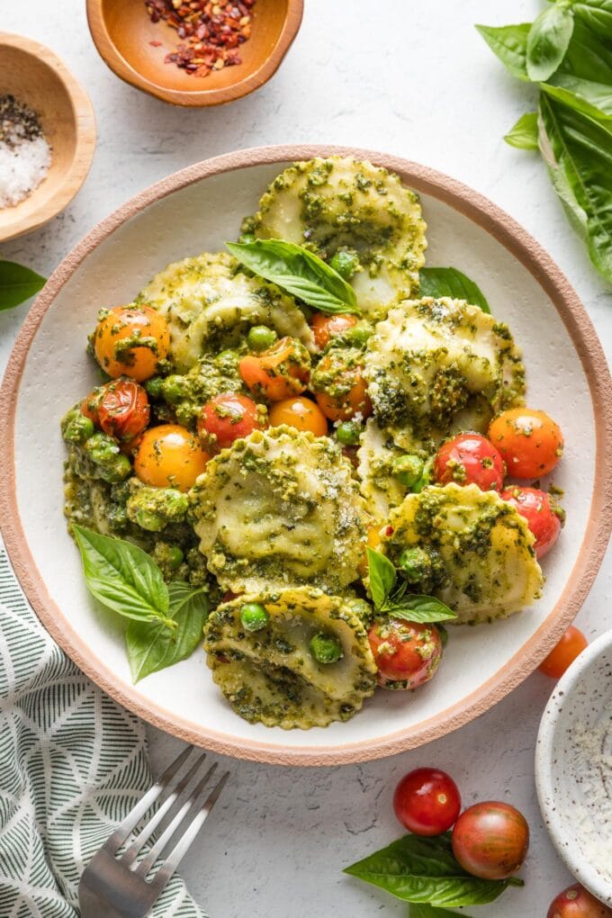 Small pasta bowl filled with pesto ravioli with peas and cherry tomatoes, with extra red pepper flakes nearby for garnish.