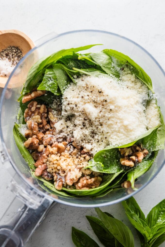 Food processor bowl filled with spinach, basil, Parmesan, walnuts, garlic, salt, and pepper.