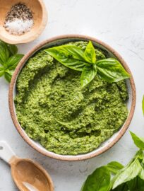 Countertop with a small bowl of basil pesto with spinach, with extra basil leaves, salt, and pepper scattered nearby.
