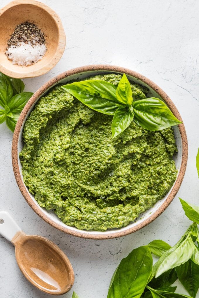 Countertop with a small bowl of basil pesto with spinach, with extra basil leaves, salt, and pepper scattered nearby.