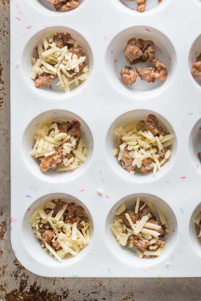 Silicone muffin tin wells filled with browned ground sausage and shredded cheddar cheese.