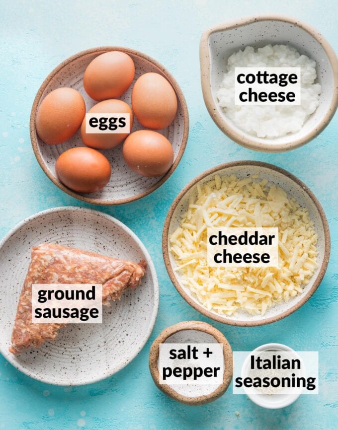 Labeled overhead photo of eggs, cottage cheese, shredded cheddar cheese, ground Italian sausage, Italian seasoning, salt, and pepper, all on plates or in prep bowls and ready to cook.