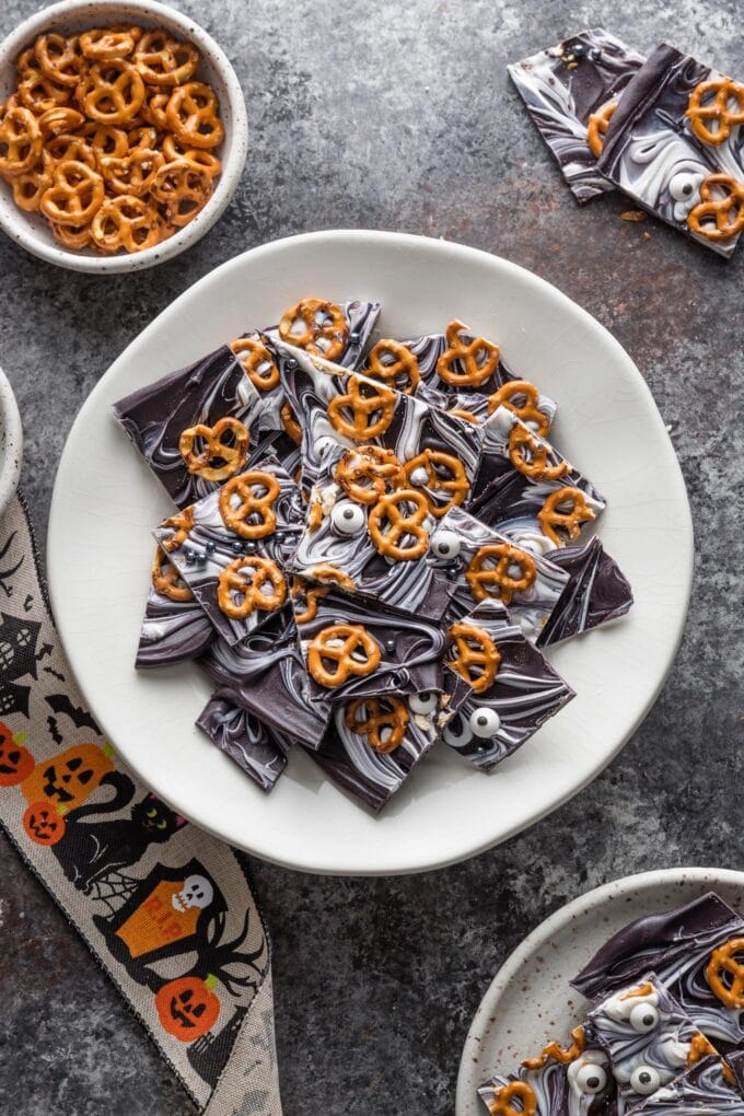 Small white plate piled high with pieces of "boo bark" -- chocolate bark with white swirls and pressed with pretzels and candy eyeballs for a Halloween treat.