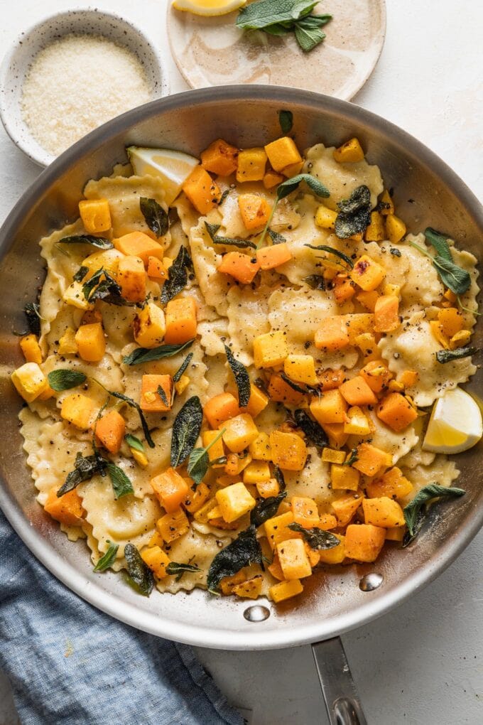 Overhead view of a skillet with ravioli in a brown butter sage sauce with roasted butternut squash cubes, black pepper, and Parmesan cheese.