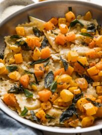 Angled view of a brown butter ravioli skillet with lemon wedges, sage leaves, and butternut squash.