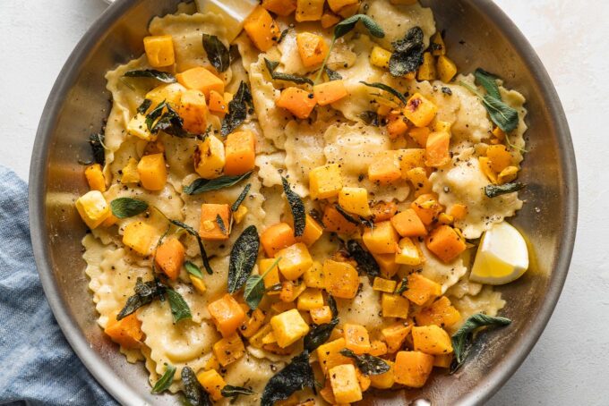Landscape oriented photo of a skillet with ravioli in a brown butter sage sauce with roasted butternut squash cubes, black pepper, and Parmesan cheese.