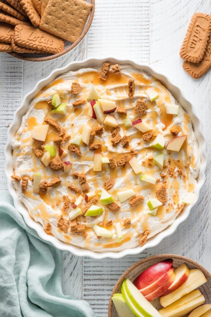 Overhead view of a shareable dessert of caramel apple inspired cheesecake dip scattered with extra apples and cookies in a flat dish.