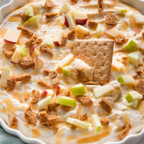 Large fluted serving dish full of creamy caramel apple cheesecake dip with a graham cracker being dipped in for a snack.