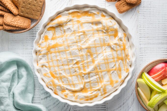 Caramel apple cheesecake dip ready to serve with extra caramel sauce drizzled on top and bowls of graham crackers and apple slices arranged nearby.