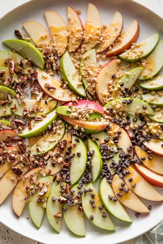 Large white platter filled with sliced red and green apples drizzled with caramel sauce and sprinkled with sea salt, toffee bits, chopped pecans, and miniature chocolate chips.