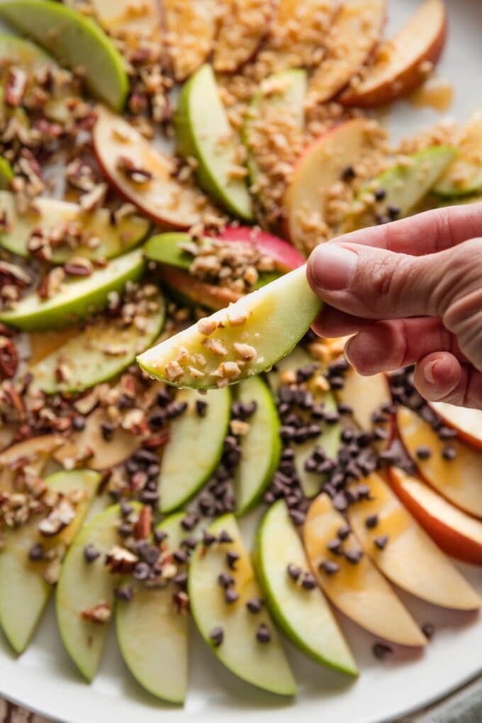 Close up of a hand holding an apple slice drizzled with caramel sauce and topped with candy toffee bits.