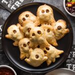Black plate loaded with ghost-shaped miniature taco pies with two types of salsa nearby for dipping.