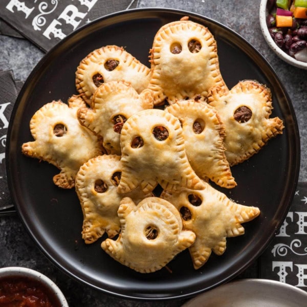 Black plate loaded with ghost-shaped miniature taco pies with two types of salsa nearby for dipping.