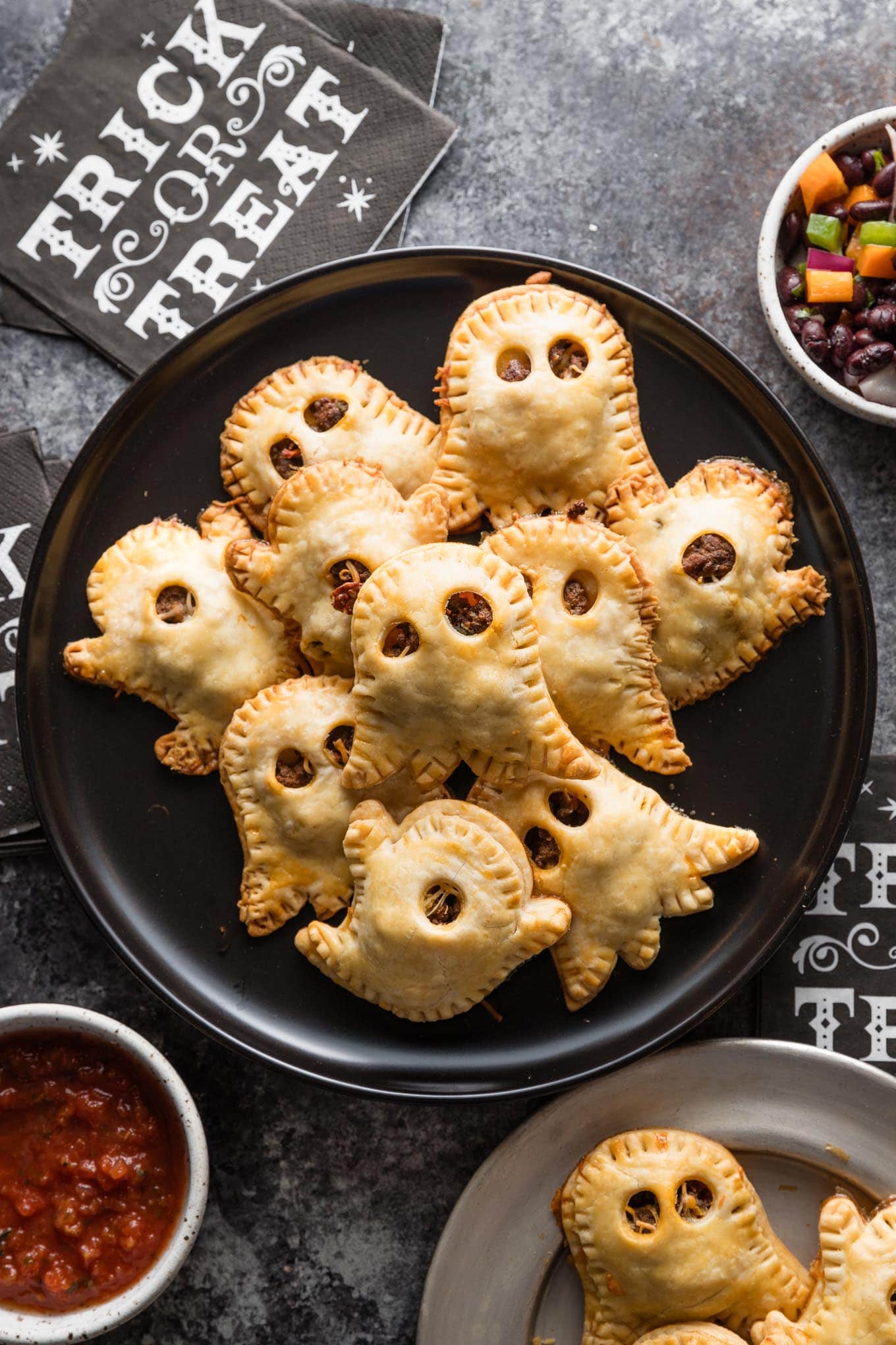 Black plate loaded with ghost-shaped miniature taco pies with two types of salsa nearby for dipping.