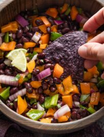 Close up of a blue corn tortilla chip scooping up a portion of a Halloween-themed black bean dip, with purple-ish red onion and green and orange bell peppers.