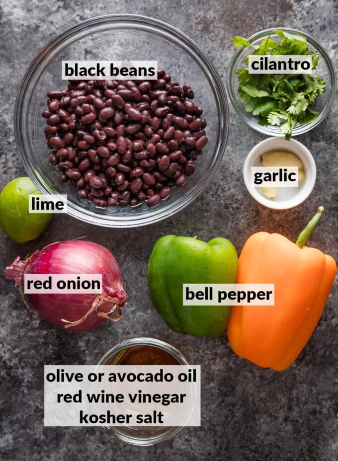 Labeled overhead photo of drained and rinsed black beans, an orange and a green bell pepper, fresh cilantro leaves, garlic cloves, a whole lime, red onion, and a small jar holding olive oil, red wine vinegar, and a pinch of kosher salt.