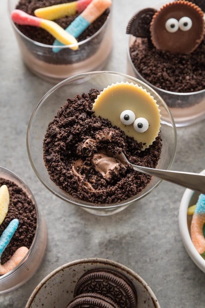 A cocktail glass filled with a Halloween-themed dirt cup dessert, with chocolate pudding, crushed Oreo toppings, and a white chocolate peanut butter cup dotted with candy eyeballs on top.