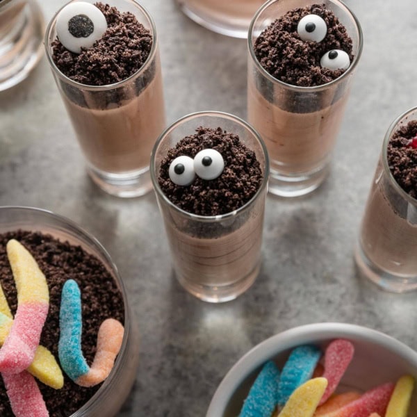 Small, tall shooter-style glasses filled with chocolate pudding and topped with crushed Oreos and candy eyeballs to make Halloween-inspired dirt cup shooters.