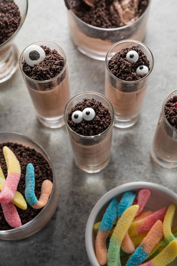 Small, tall shooter-style glasses filled with chocolate pudding and topped with crushed Oreos and candy eyeballs to make Halloween-inspired dirt cup shooters.