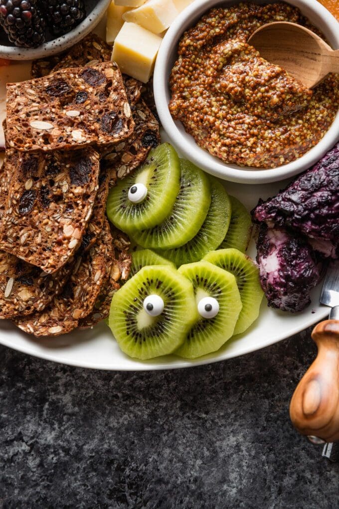 Slices of green kiwi fruit decorated with candy eyeballs to look like "monsters" on a Halloween fruit and cheese board.