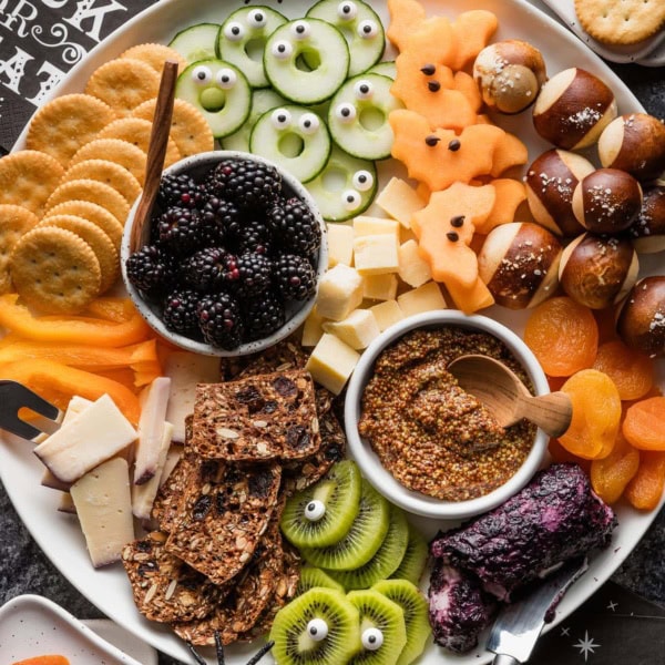 Overhead view of a large cheese board with a Halloween theme including one-eyed kiwi slices, cantaloupe bats, monster cucumber slices, and more.