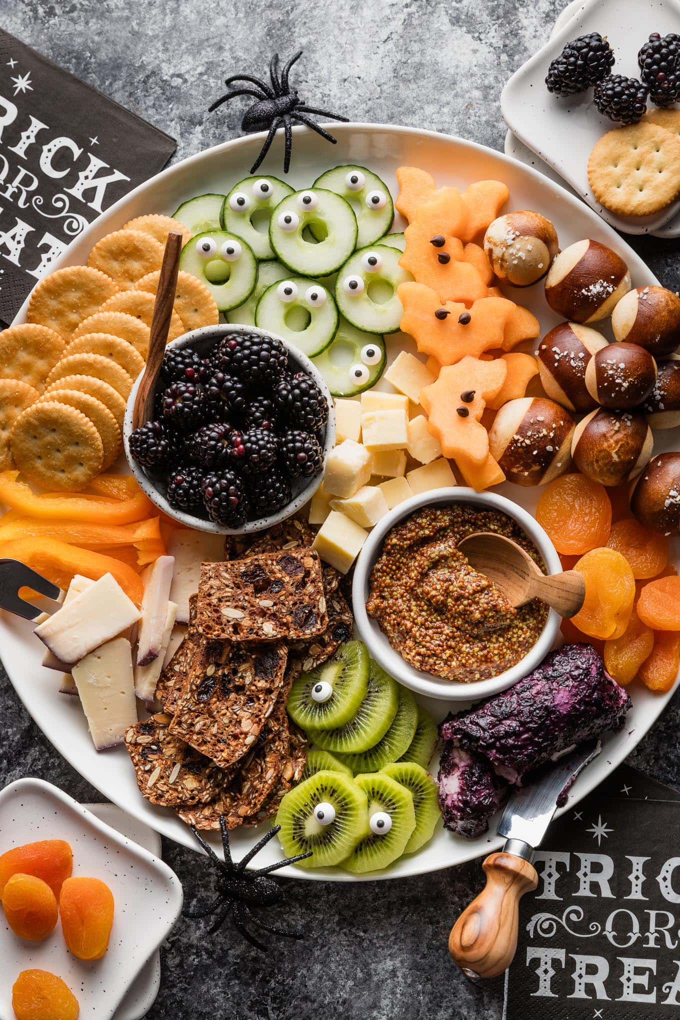 Overhead view of a large cheese board with a Halloween theme including one-eyed kiwi slices, cantaloupe bats, monster cucumber slices, and more.