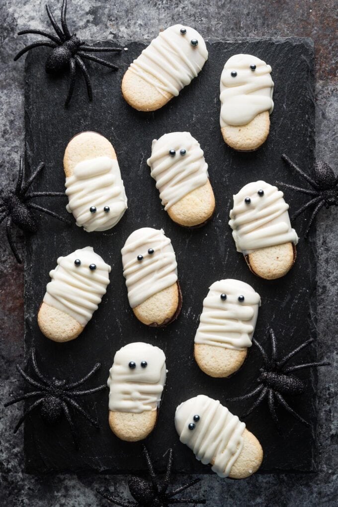 Chalkboard style tray set on a dark gray countertop and scattered with Milano mummy cookies and black sparkly decorative spiders.