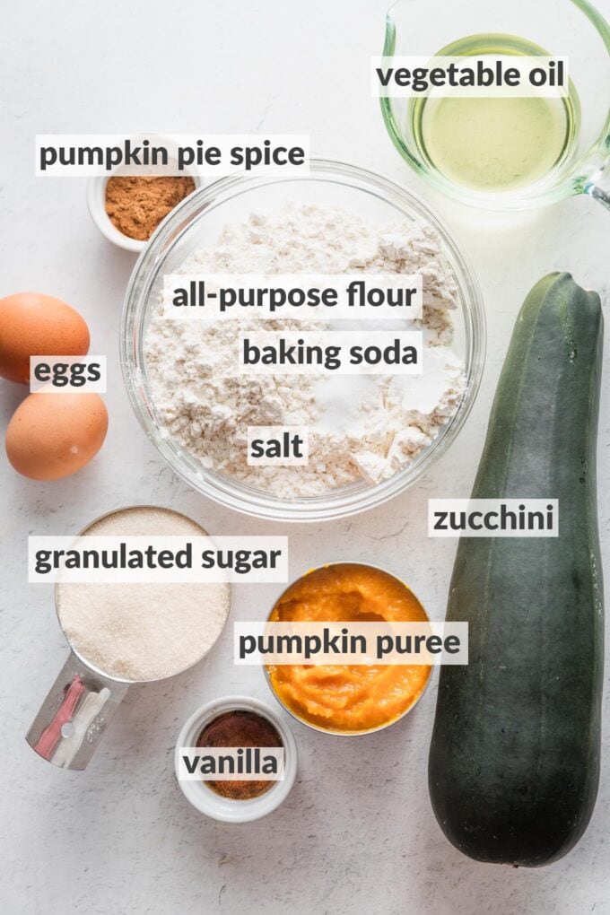 Labeled overhead photo of all-purpose flour, baking soda, salt, a whole zucchini, an open can of pumpkin puree, vanilla extract, vegetable oil, granulated sugar, two eggs, and pumpkin pie spice,  in prep bowls and ready to bake.