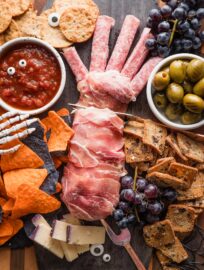 Cheese and cracker board designed for Halloween, with a prosciutto-wrapped cheese log made to look like a cut off hand, black and orange tortilla chips, black grapes, grilled olives, and candy eyes.
