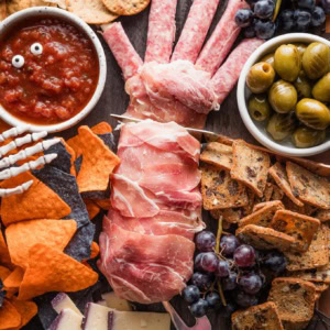 Cheese and cracker board designed for Halloween, with a prosciutto-wrapped cheese log made to look like a cut off hand, black and orange tortilla chips, black grapes, grilled olives, and candy eyes.