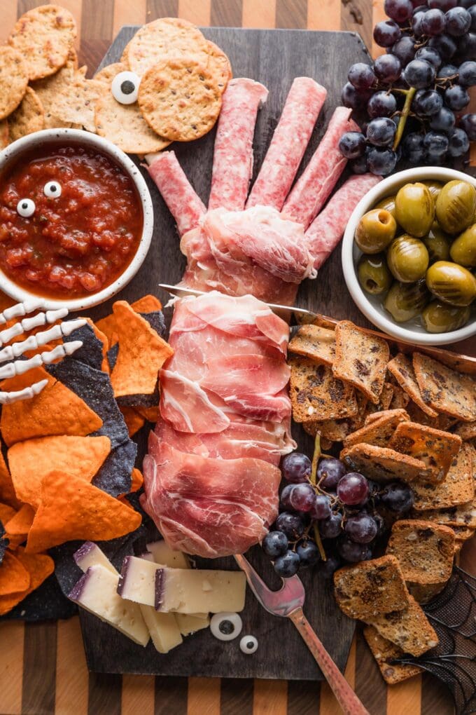 Cheese and cracker board designed for Halloween, with a prosciutto-wrapped cheese log made to look like a cut off hand, black and orange tortilla chips, black grapes, grilled olives, and candy eyes.