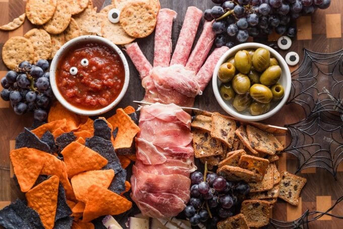 Landscape view of a Halloween-inspired cheeseboard with a prosciutto-wrapped cheese log sliced off to resemble a disembodied hand.
