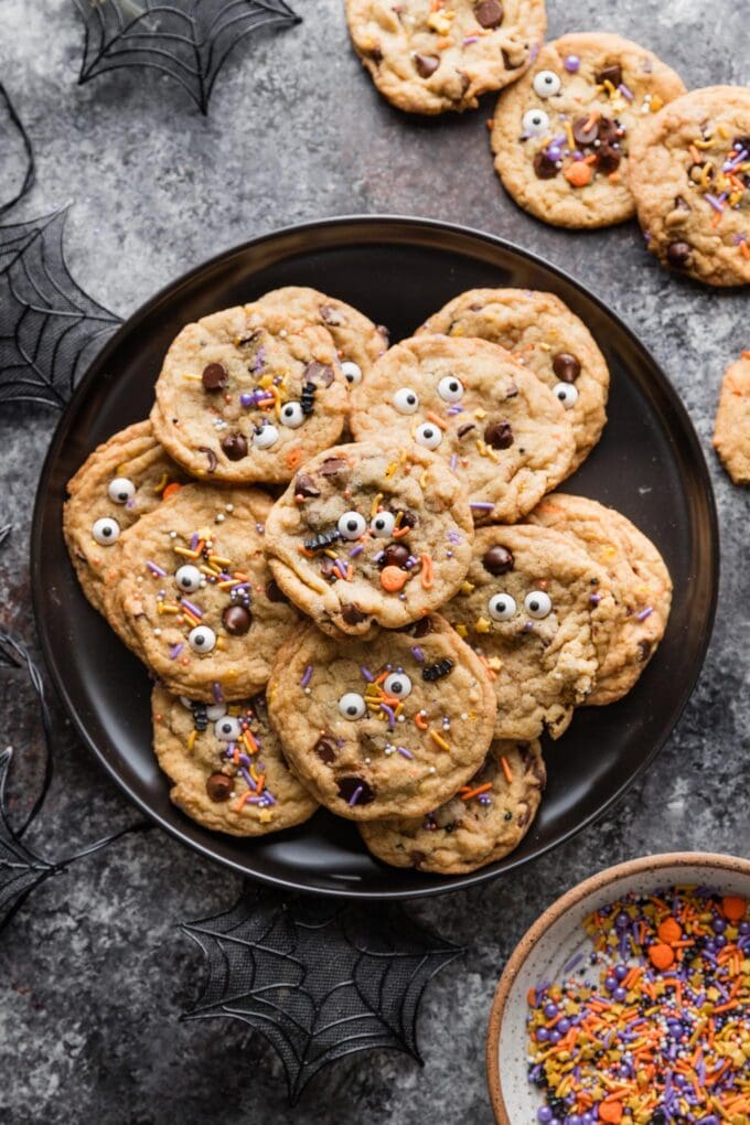 Dark countertop with a plate of spooky themed chocolate chip cookies, extra bat and pumpkin sprinkles, and a black spiderweb decoration.