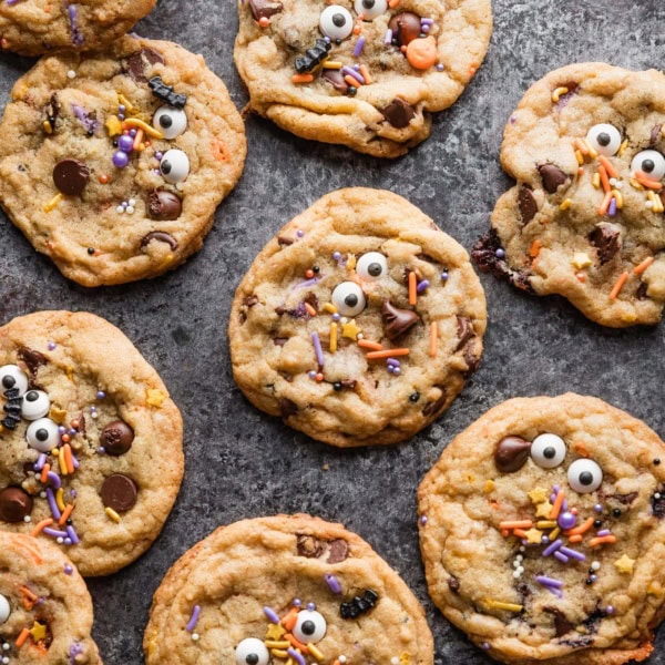 Dark gray countertop scattered with chocolate chip cookies decorated with candy eyes and Halloween bat and pumpkin themed sprinkles.