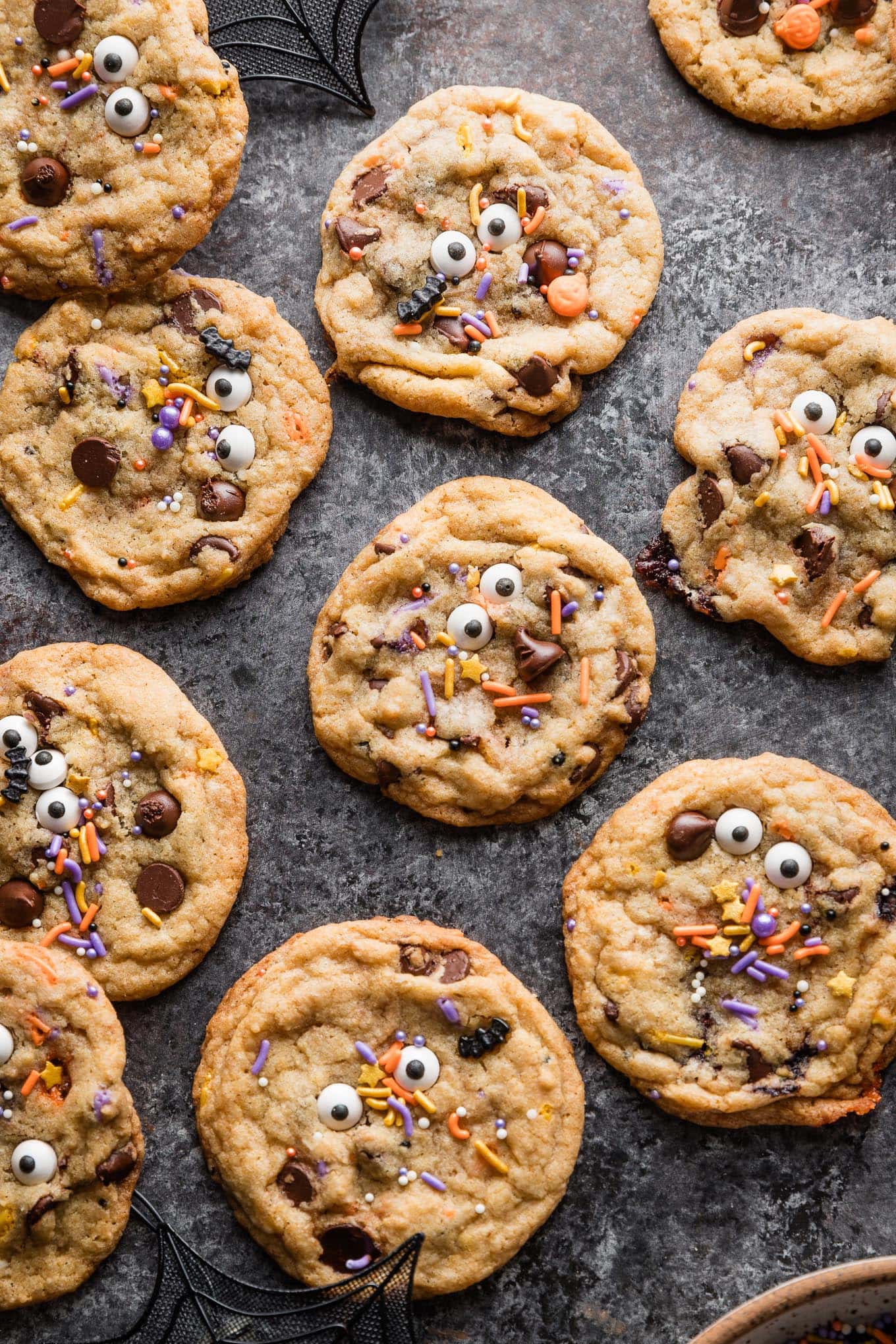 Dark gray countertop scattered with chocolate chip cookies decorated with candy eyes and Halloween bat and pumpkin themed sprinkles.