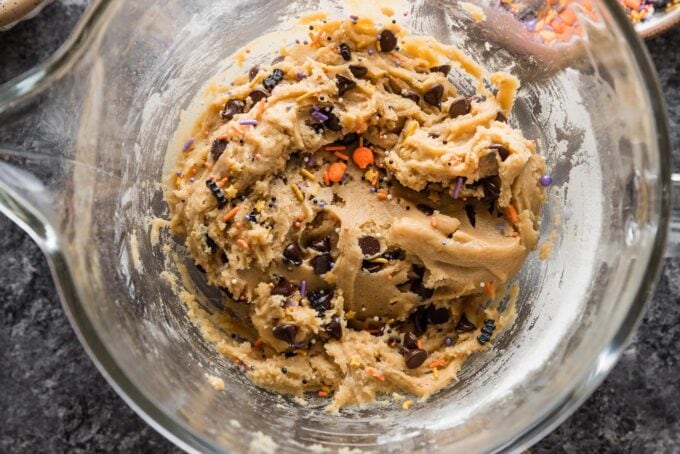 Close up of chocolate chip cookie dough with Halloween sprinkles mixed in in a clear glass stand mixer bowl.