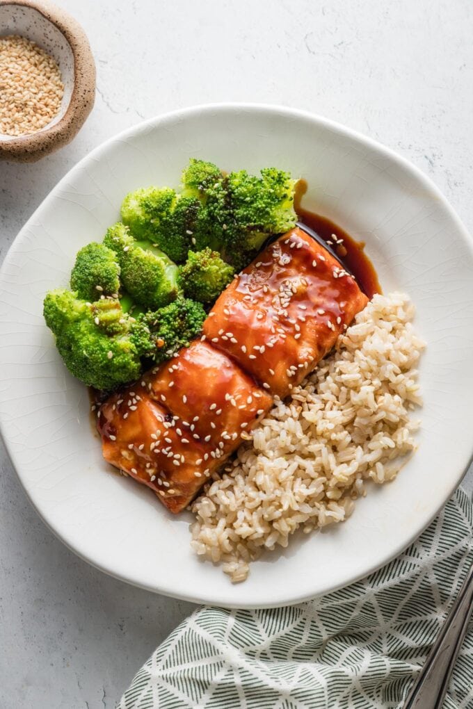 Countertop set with a plate of teriyaki salmon, steamed broccoli, and brown rice, with a patterned napkin and a bowl of sesame seeds for garnish set nearby.