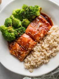 Small white plate with a meal of teriyaki coated salmon garnished with sesame seeds, brown rice, and steamed broccoli.