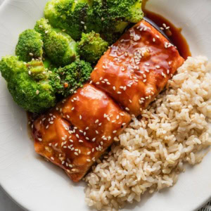 Small white plate with a meal of teriyaki coated salmon garnished with sesame seeds, brown rice, and steamed broccoli.