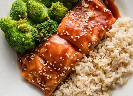 Small white plate with a meal of teriyaki coated salmon garnished with sesame seeds, brown rice, and steamed broccoli.
