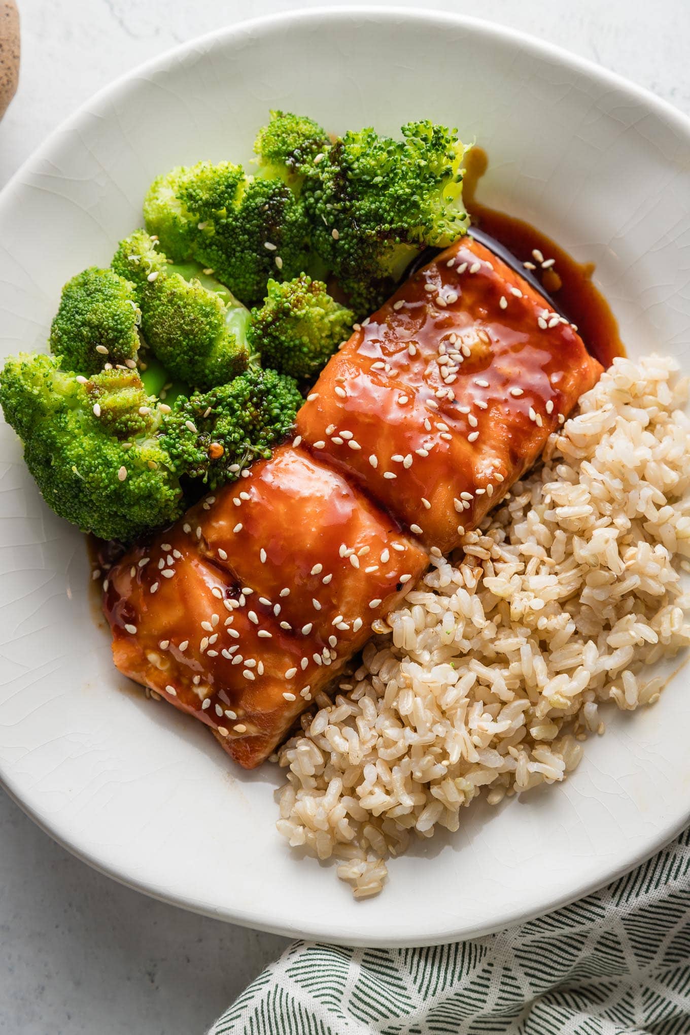 Small white plate with a meal of teriyaki coated salmon garnished with sesame seeds, brown rice, and steamed broccoli.
