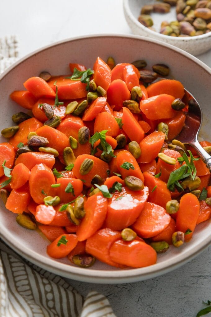 Angled view of glazed carrots with parsley and pistachios in a small serving bowl.