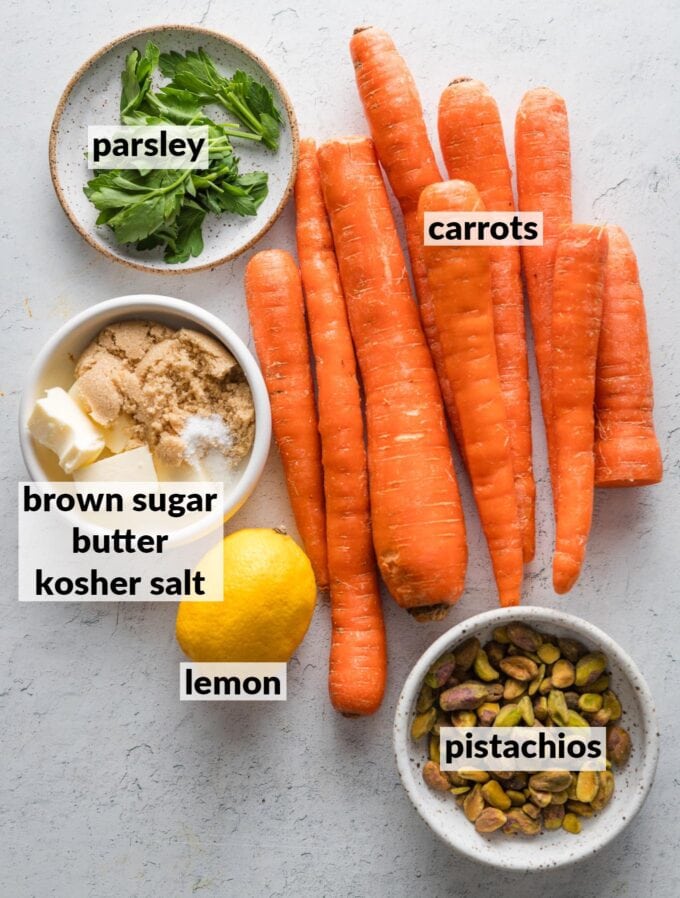 Labeled overhead photo of whole carrots, fresh parsley, pistachio nutmeats, a whole lemon, and a small bowl with butter, brown sugar, and salt.