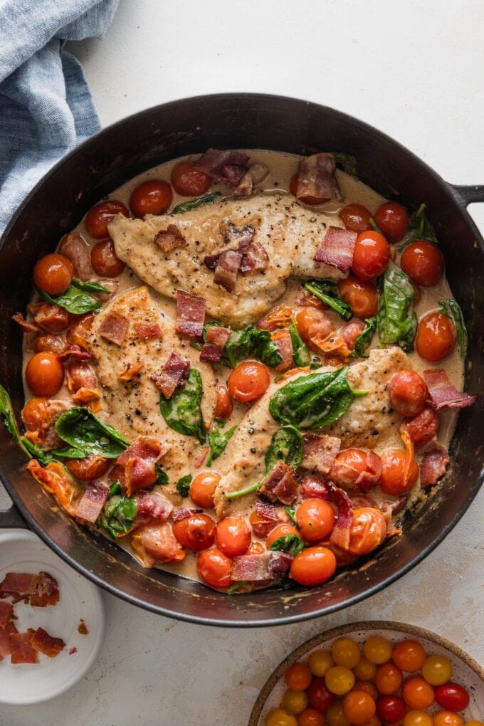 Overhead view of a large cast iron skillet with a dish of pan seared chicken breasts with bacon and cherry tomatoes in a balsamic cream sauce with baby spinach. 