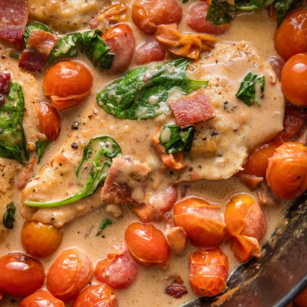 Close up of thin sliced chicken breasts cooked in a skillet with bacon, a light balsamic cream sauce, baby spinach, and cherry tomatoes.