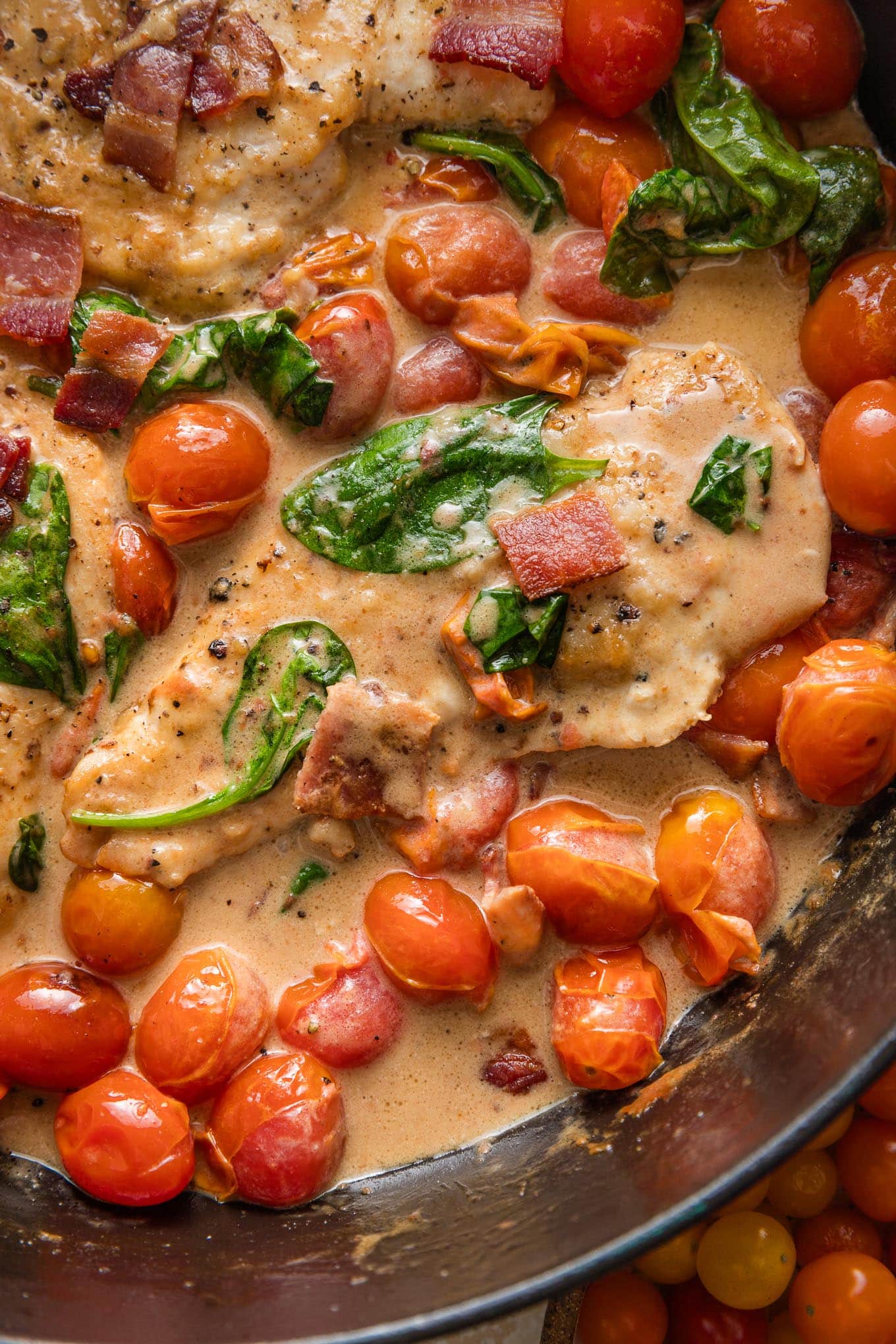 Close up of thin sliced chicken breasts cooked in a skillet with bacon, a light balsamic cream sauce, baby spinach, and cherry tomatoes.