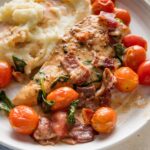 White plate filled with a helping of chicken with bacon in a balsamic cream sauce with cherry tomatoes and a side of mashed potatoes.