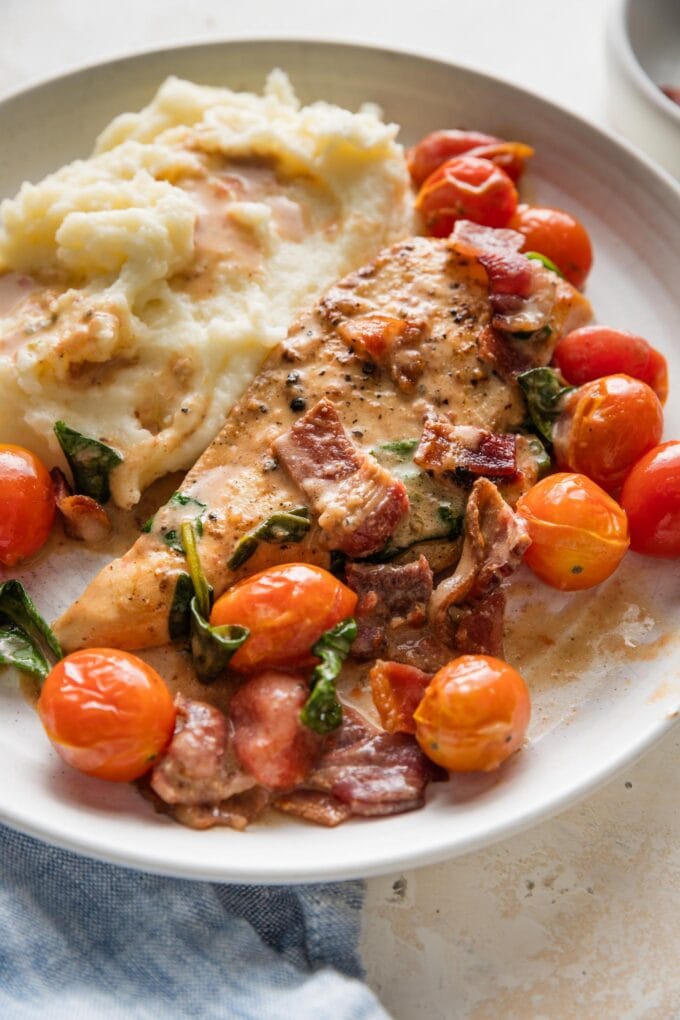 White plate filled with a helping of chicken with bacon in a balsamic cream sauce with cherry tomatoes and a side of mashed potatoes.