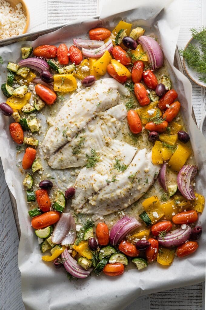 Large sheet pan with baked tilapia and lots of vegetables seasoned with Greek flavors.