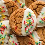 Close up of a ginger molasses cookie with a deep crackly top partially dipped in white chocolate and finished with festive Christmas sprinkles.